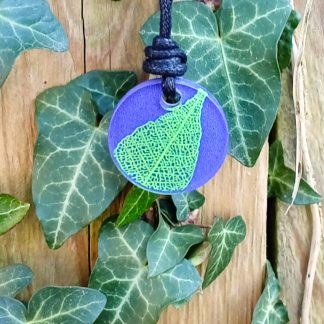 Deep purple round pendant with bright green leaf skeleton design against ivy covered wood in natural daylight